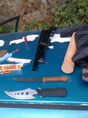 Photograph of a shotgun, two knives, a shot gun shell, and needles on the roof of a car.