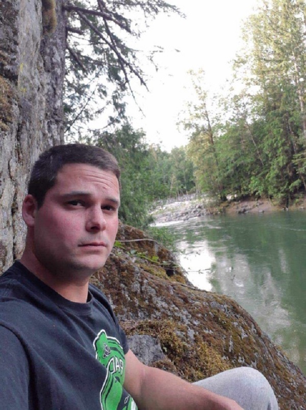 Homme des Premières Nations portant un tee-shirt foncé garni d’un logo vert. L’homme est assis dehors, près d’une rivière. 